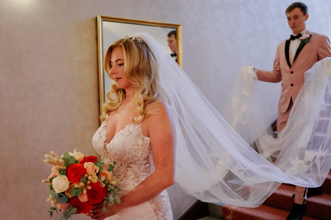 A bride holding a bouquet stands with her eyes closed while a man in a suit holds her veil on a staircase.