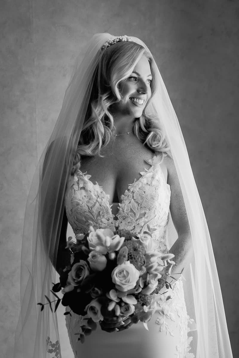 A black and white portrait of a smiling bride holding a bouquet while wearing a veil and an intricate lace dress.