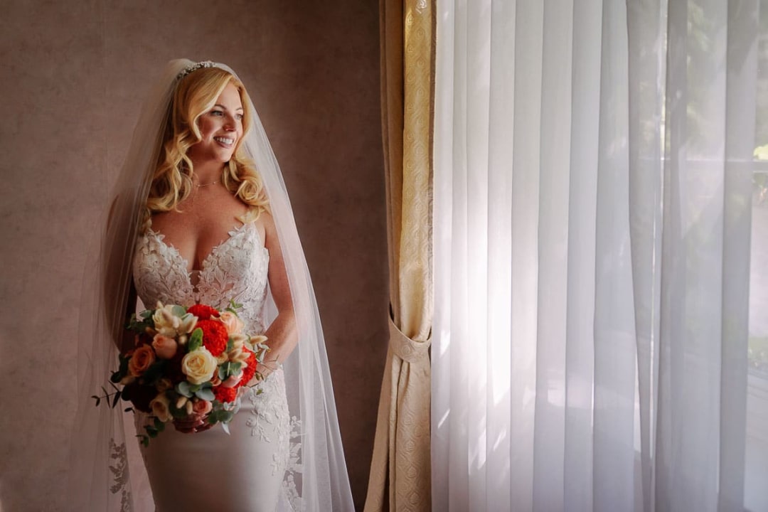A bride in an elegant lace dress and veil stands by a window, holding a vibrant bouquet and smiling softly.