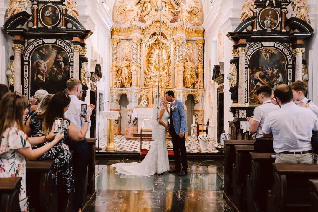 A bride and groom sharing a kiss at the altar inside a beautifully decorated church, surrounded by wedding guests taking photos.