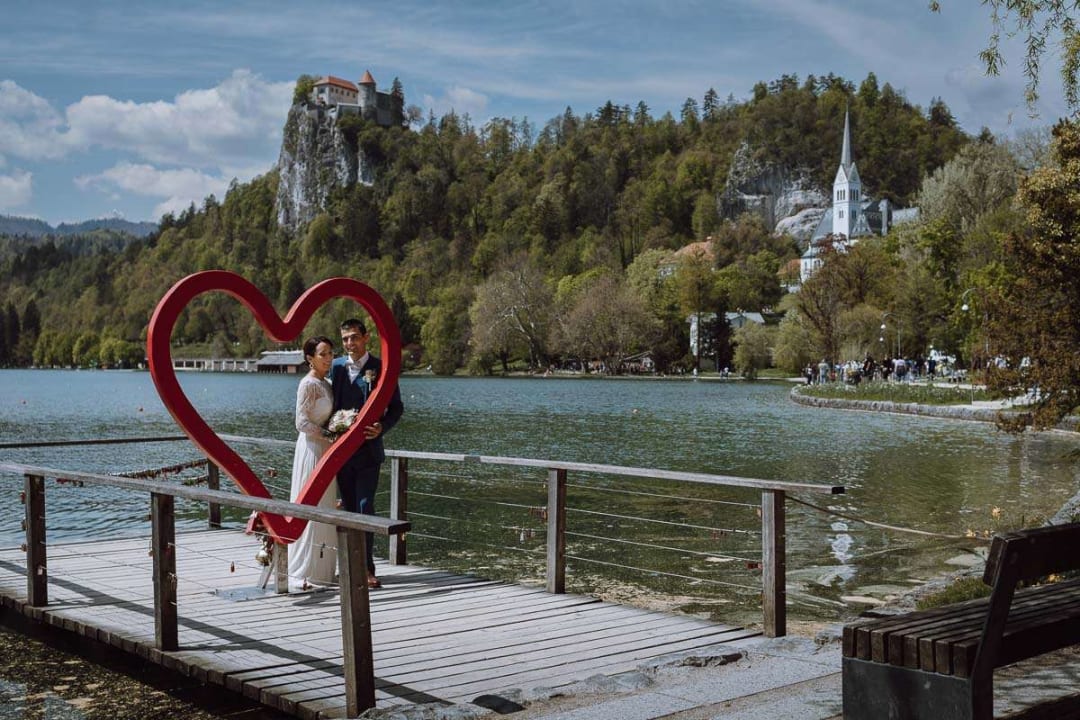 A bride and groom stand on a dock by Lake Bled, framed by a large red heart sculpture, with the Bled Castle and a church visible on the hillside in the background.