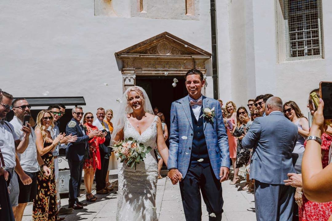 A bride and groom happily walking hand in hand through a crowd of applauding guests outside a church.