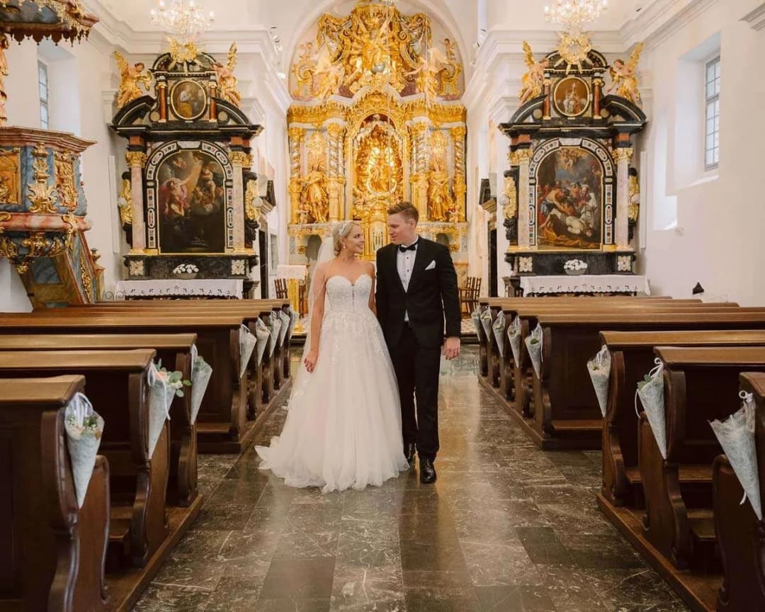 A bride and groom walking down the aisle in a beautifully decorated church with ornate gold details and paintings.