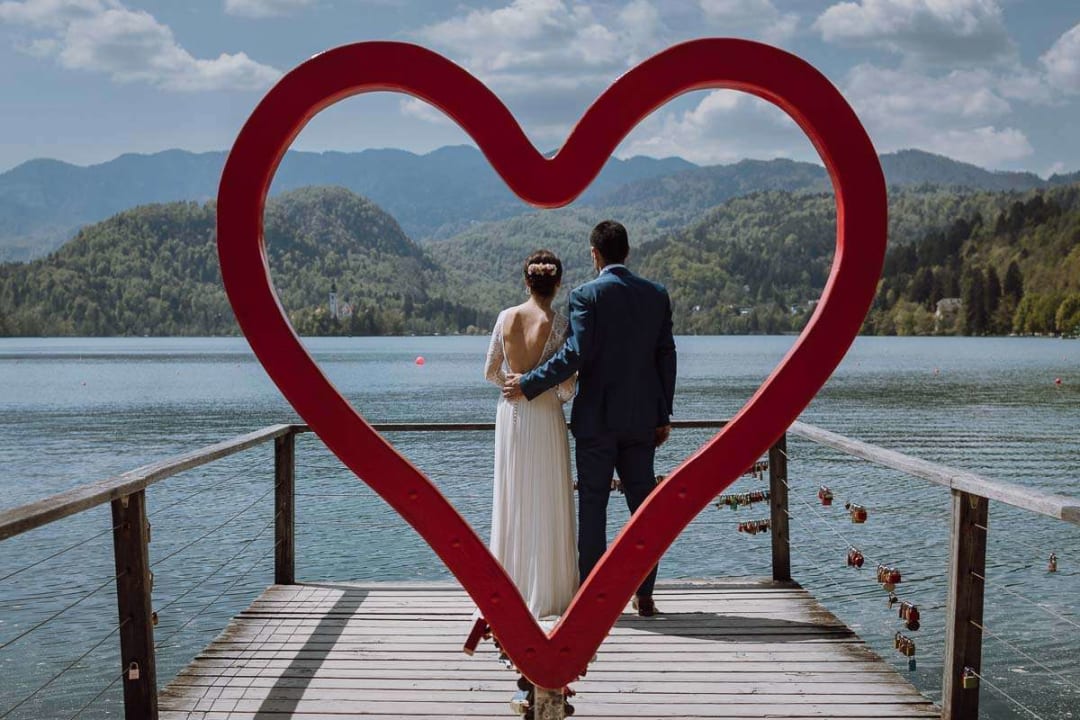 A couple stands on a dock at Lake Bled, framed by a large red heart sculpture, with mountains in the background.