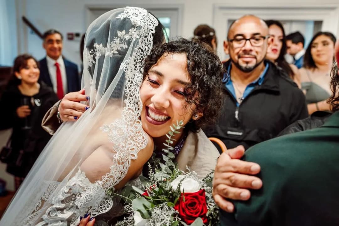 A bride in a lace veil hugging a smiling guest who is holding a bouquet of red and white roses at a wedding reception.