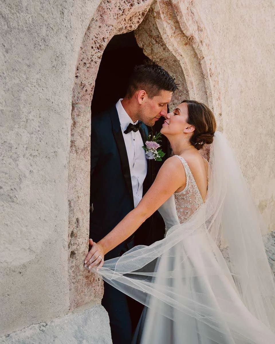 A bride and groom share a tender kiss while standing in a stone archway, with the bride's veil flowing around them.