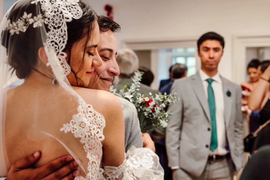 A bride and groom embracing affectionately during a wedding ceremony indoors, surrounded by guests in the background.