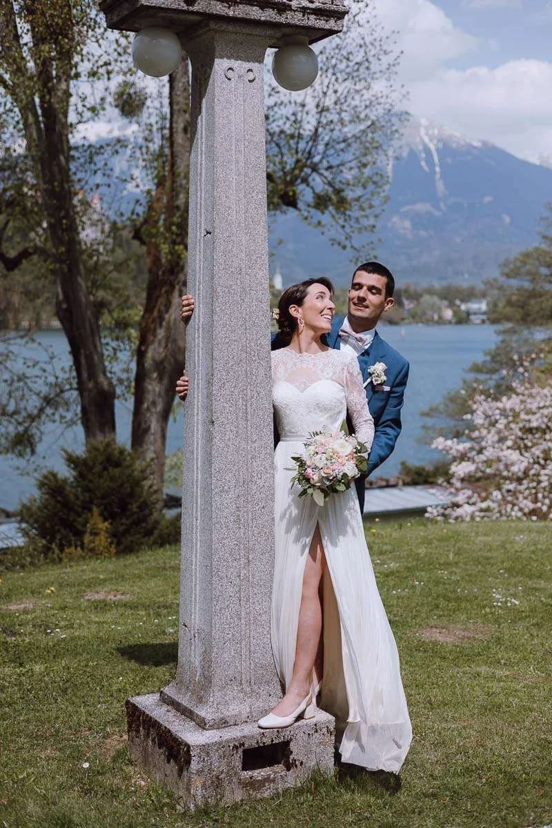 A bride and groom smiling and embracing behind a stone pillar with a view of Lake Bled and mountains in the background.