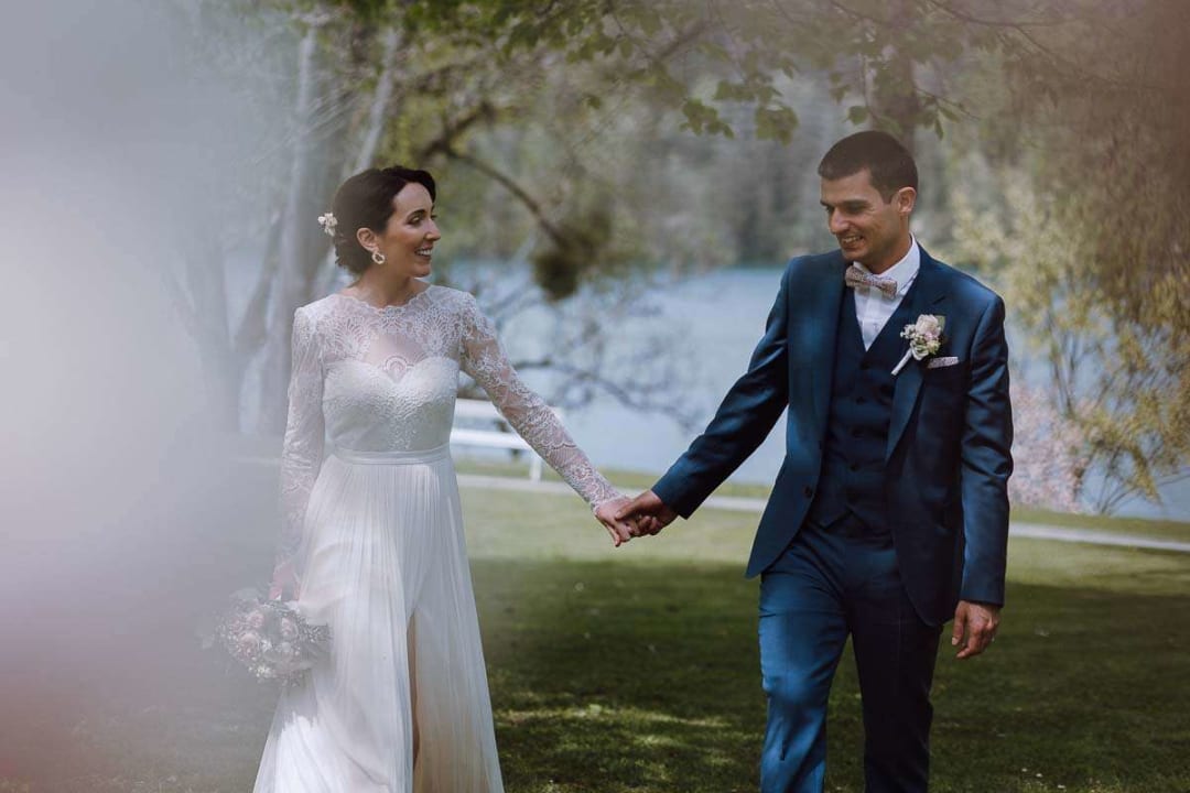 A bride and groom holding hands and smiling as they walk on the grass near a lake, surrounded by trees.
