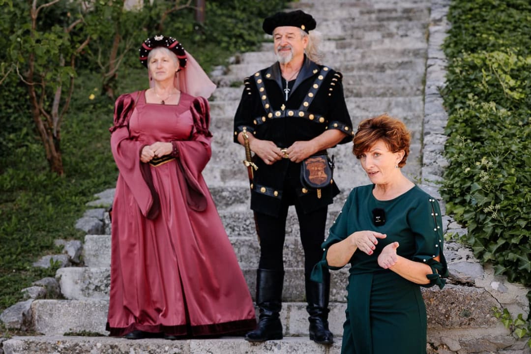 Two people in medieval attire are standing on stone steps, accompanied by a woman in a green dress, likely narrating or introducing.