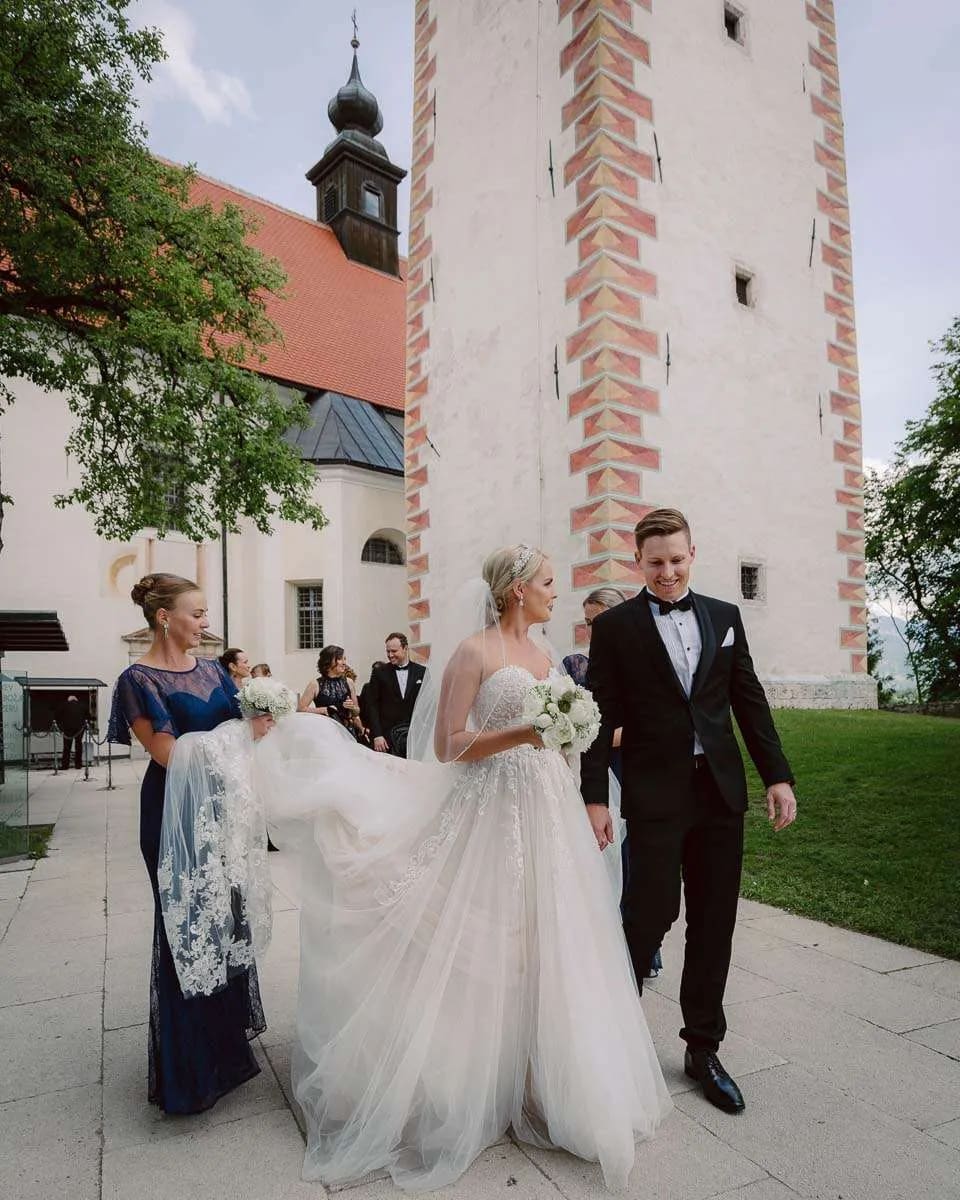A bride and groom walking happily beside a church, with the bride holding a bouquet and assisted with her train.