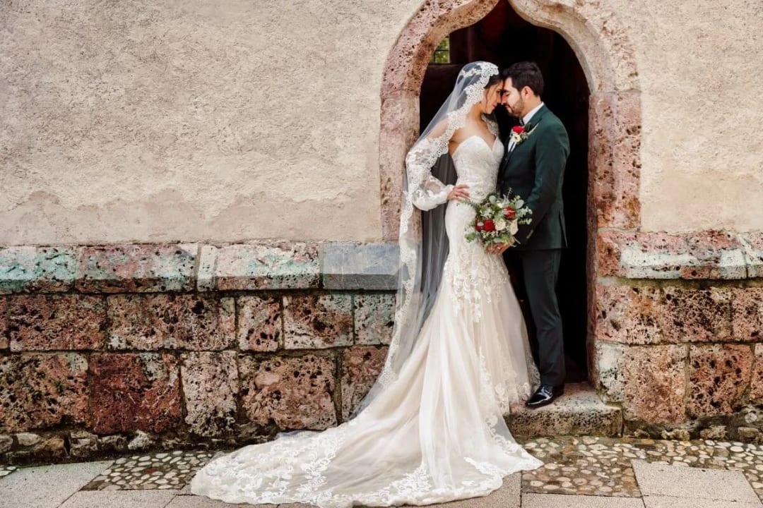 A bride and groom standing close together in a romantic pose under a stone archway, with the bride holding a bouquet of flowers.