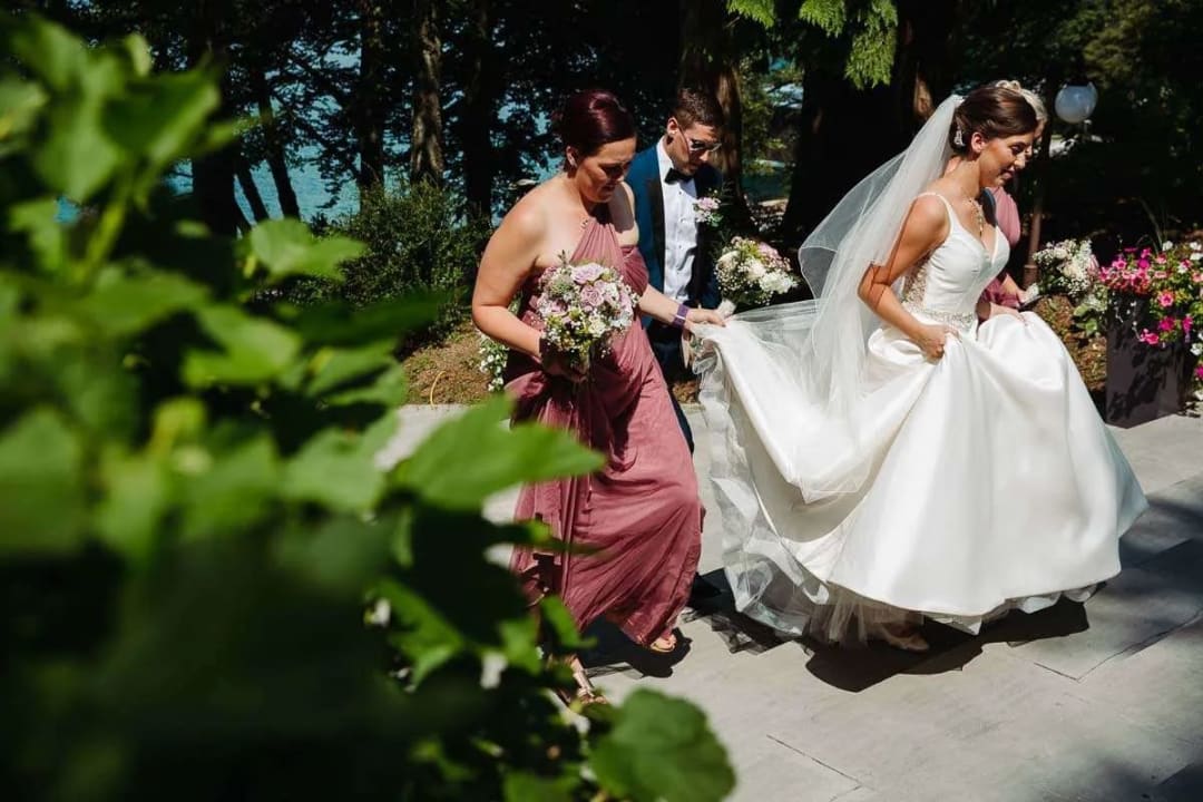 A bride in a white gown walks with her bridesmaids, holding bouquets, on a sunny day surrounded by greenery.
