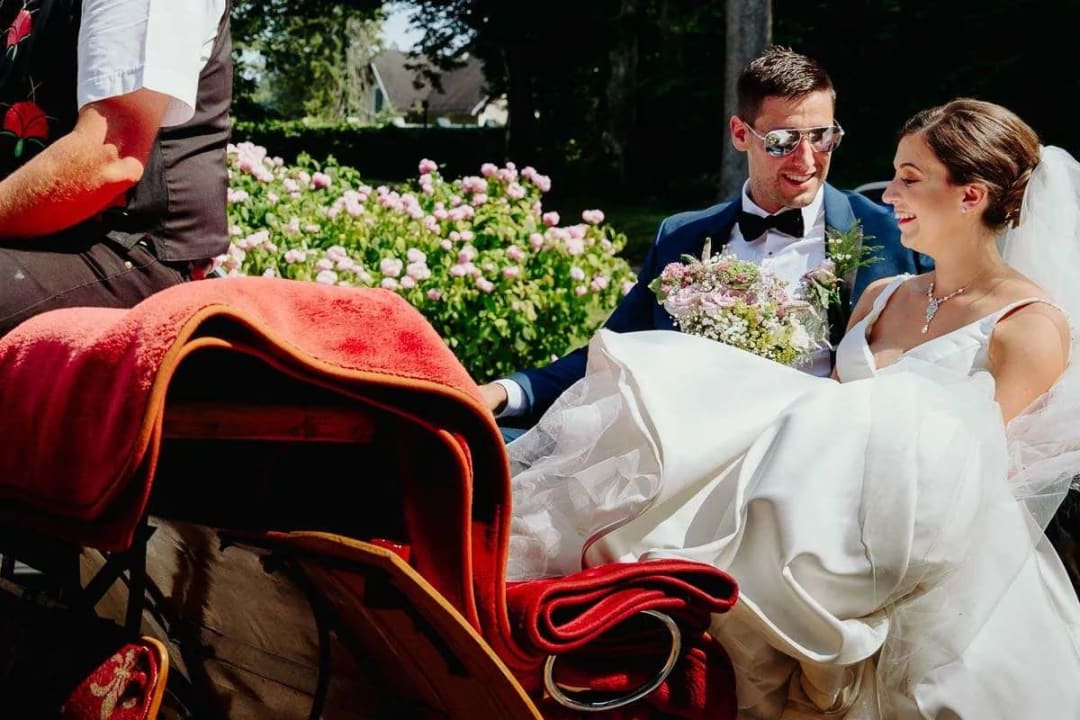 A bride and groom sitting together in a carriage surrounded by blooming pink flowers, smiling at each other.