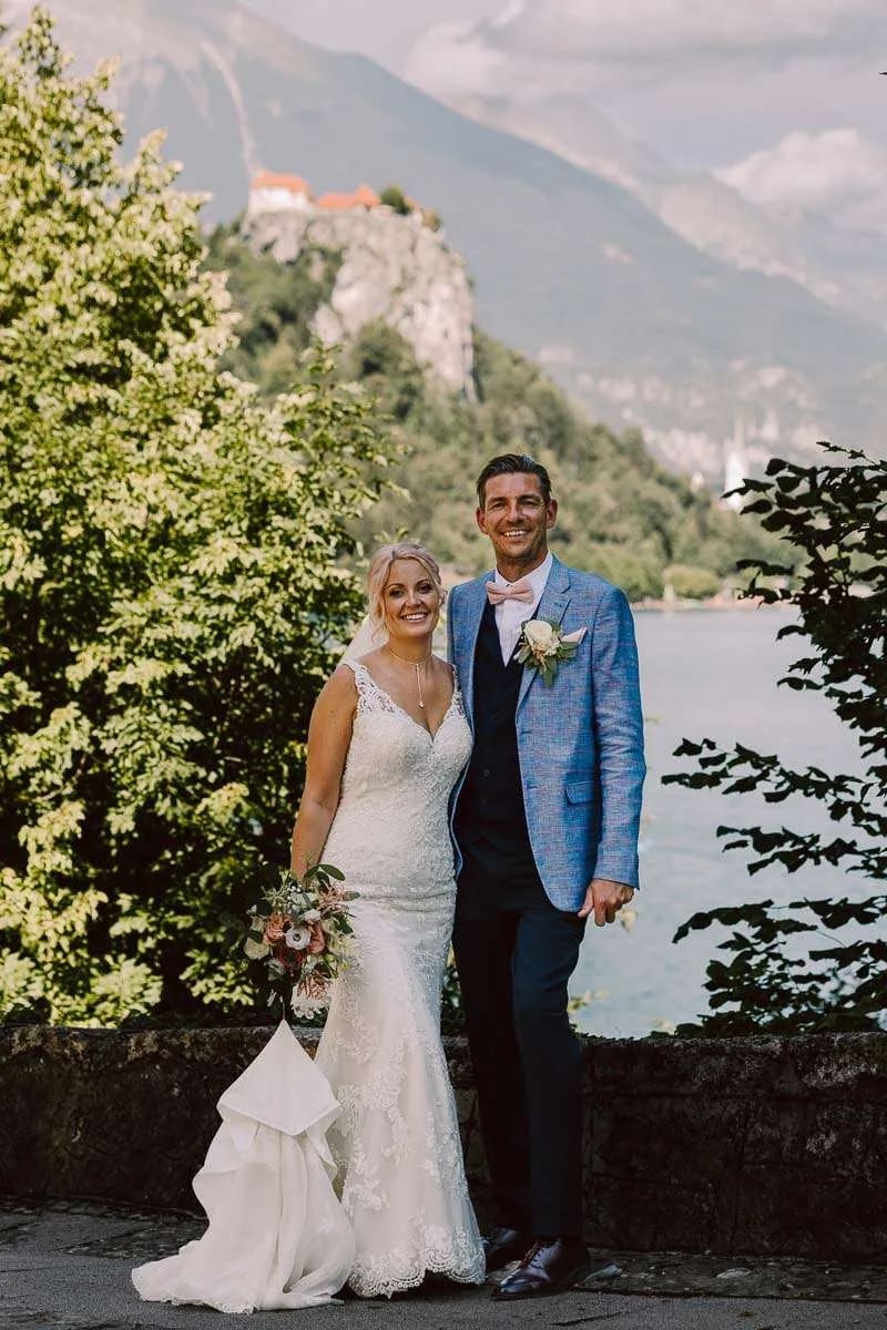 A smiling bride and groom posing together with a view of a castle on a hill and lush greenery in the background.