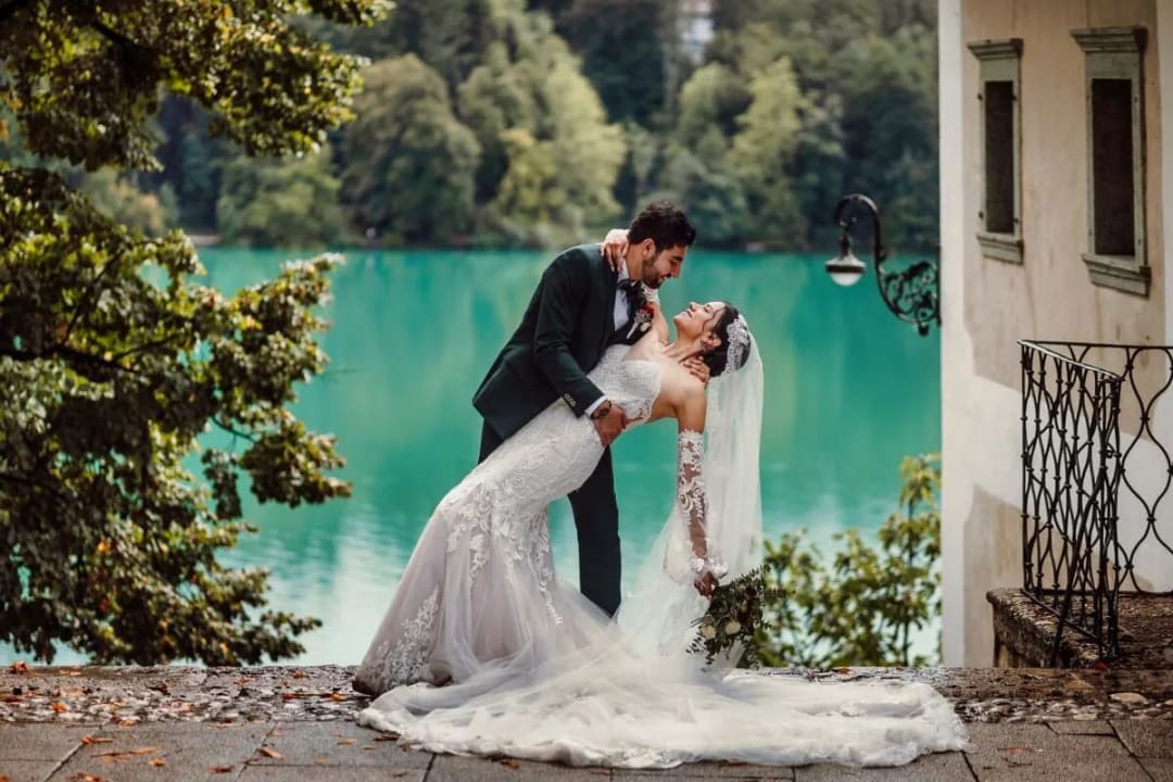 A groom affectionately dipping his bride near Lake Bled, with serene turquoise water and lush trees in the background.