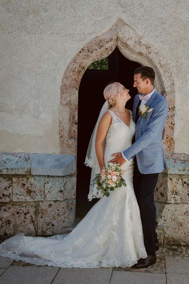 A bride and groom standing closely together, smiling at each other under an ornate stone archway.