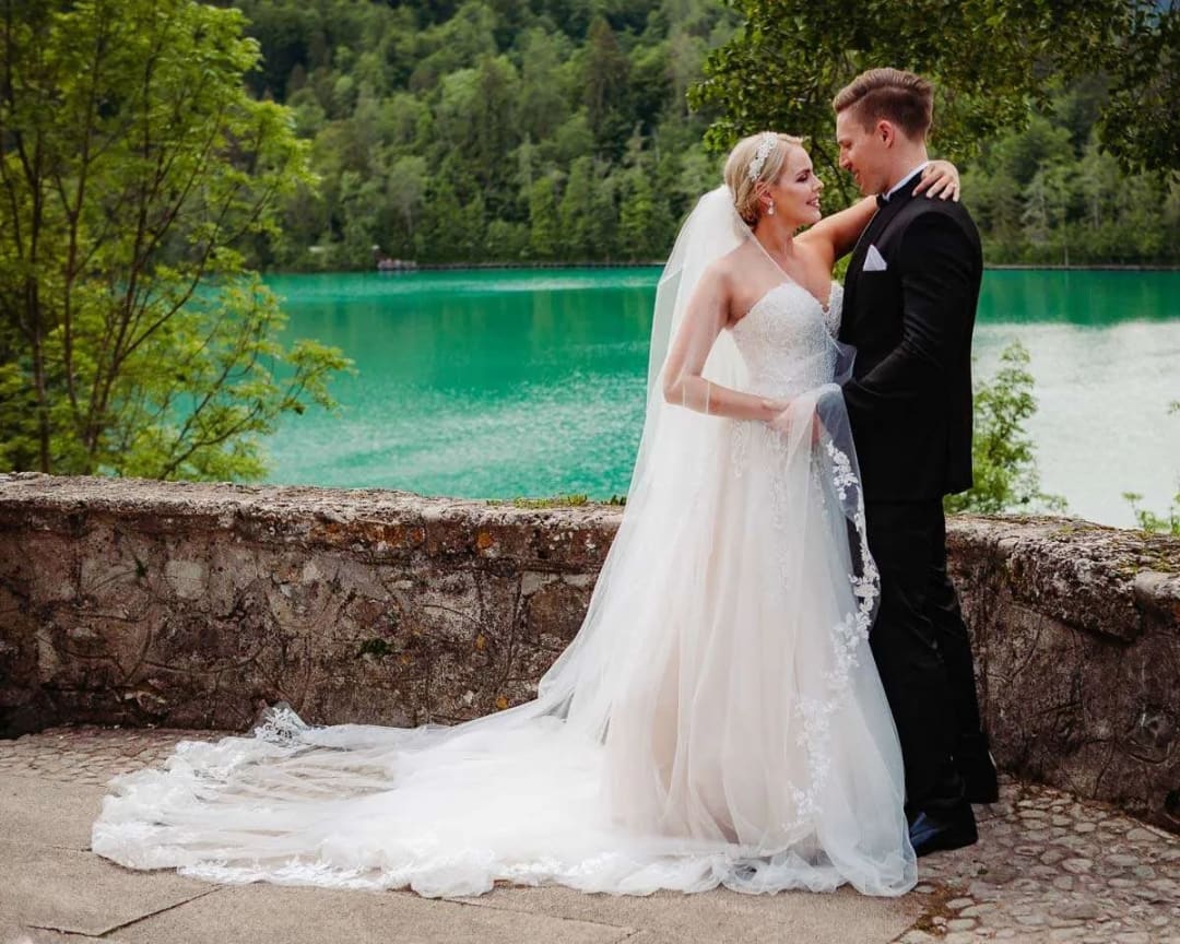A bride and groom embracing by the turquoise waters of a lake with a forested backdrop.