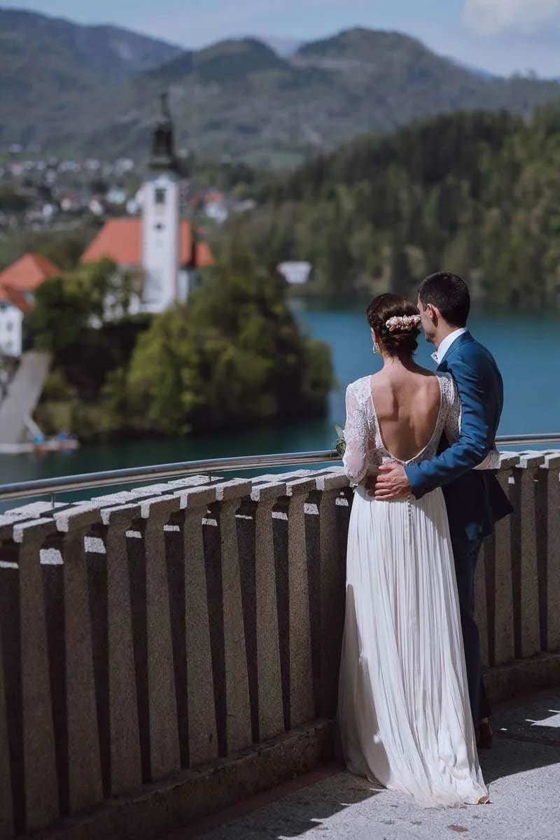 A bride and groom stand together, gazing at the scenic view of Lake Bled with the iconic church on the island and mountains in the background.