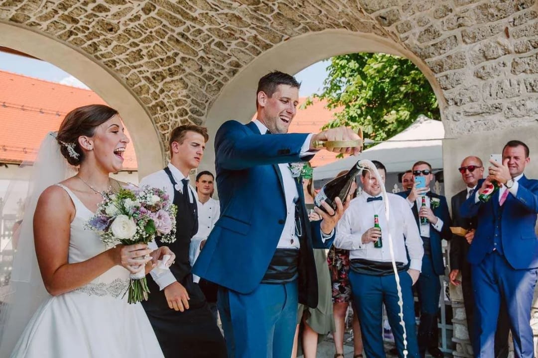 A bride and groom joyfully opening a bottle of champagne surrounded by guests at a wedding celebration.