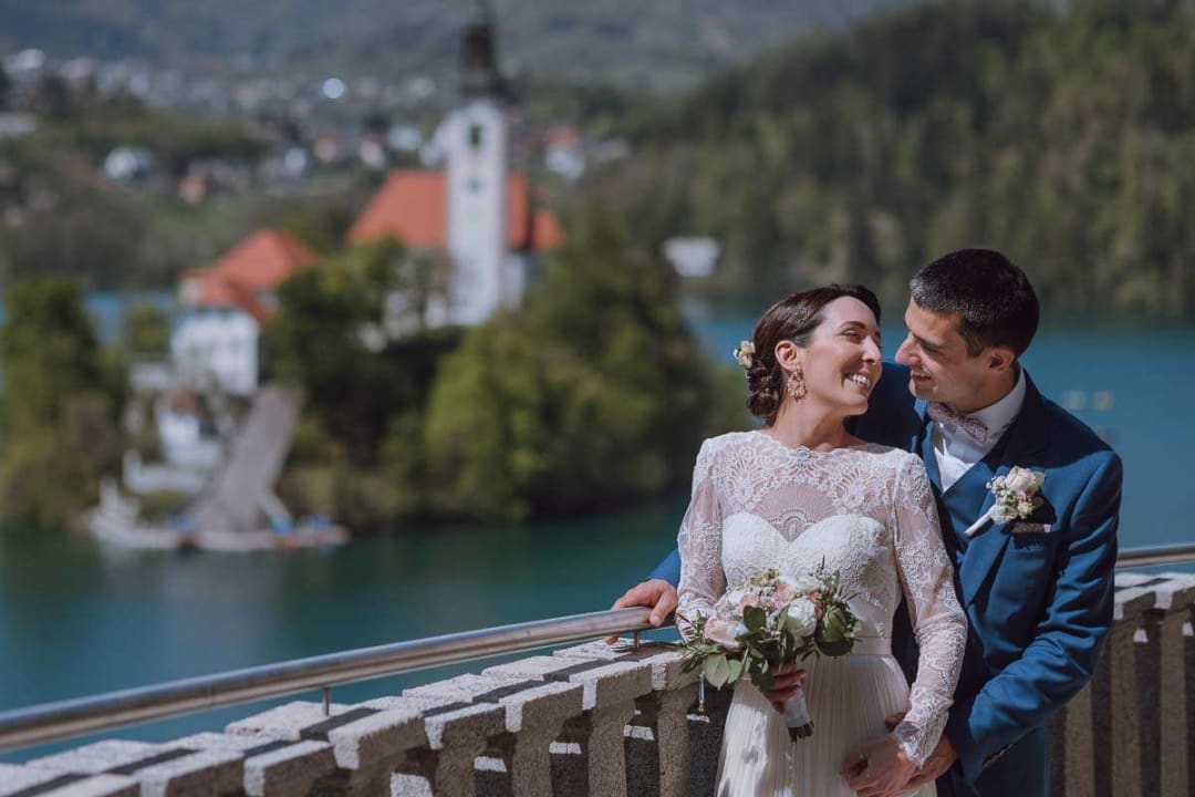 A bride and groom smiling at each other while standing on a balcony overlooking Lake Bled with a picturesque view of the church on the island in the background.