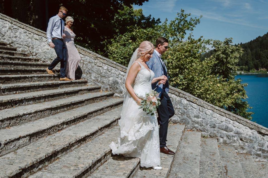 A bride and groom walking down stone steps with lush greenery and a lake in the background on a sunny day.