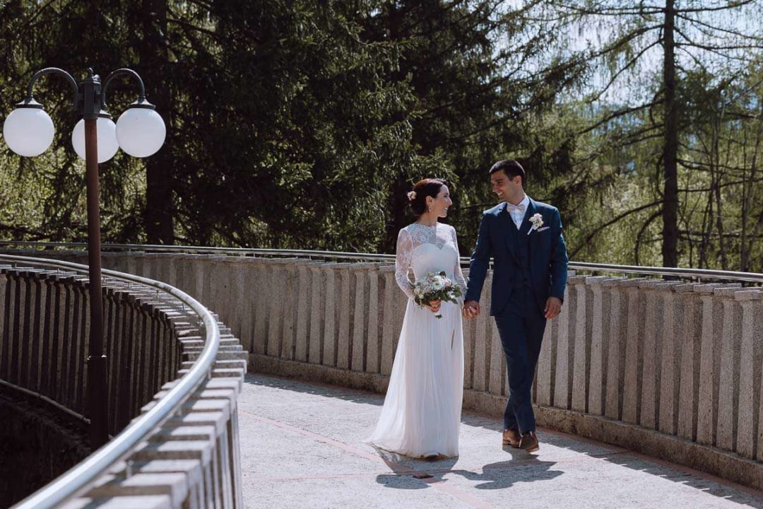 A bride and groom walking hand-in-hand along a curved pathway, surrounded by lush green trees.