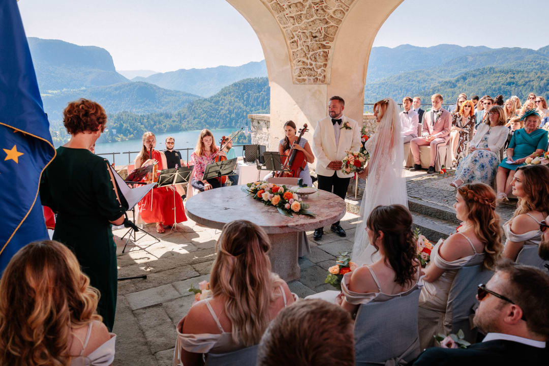 A wedding ceremony at Lake Bled, featuring a bride and groom at the altar with seated guests and a string quartet playing music, overlooking the scenic mountains and lake.