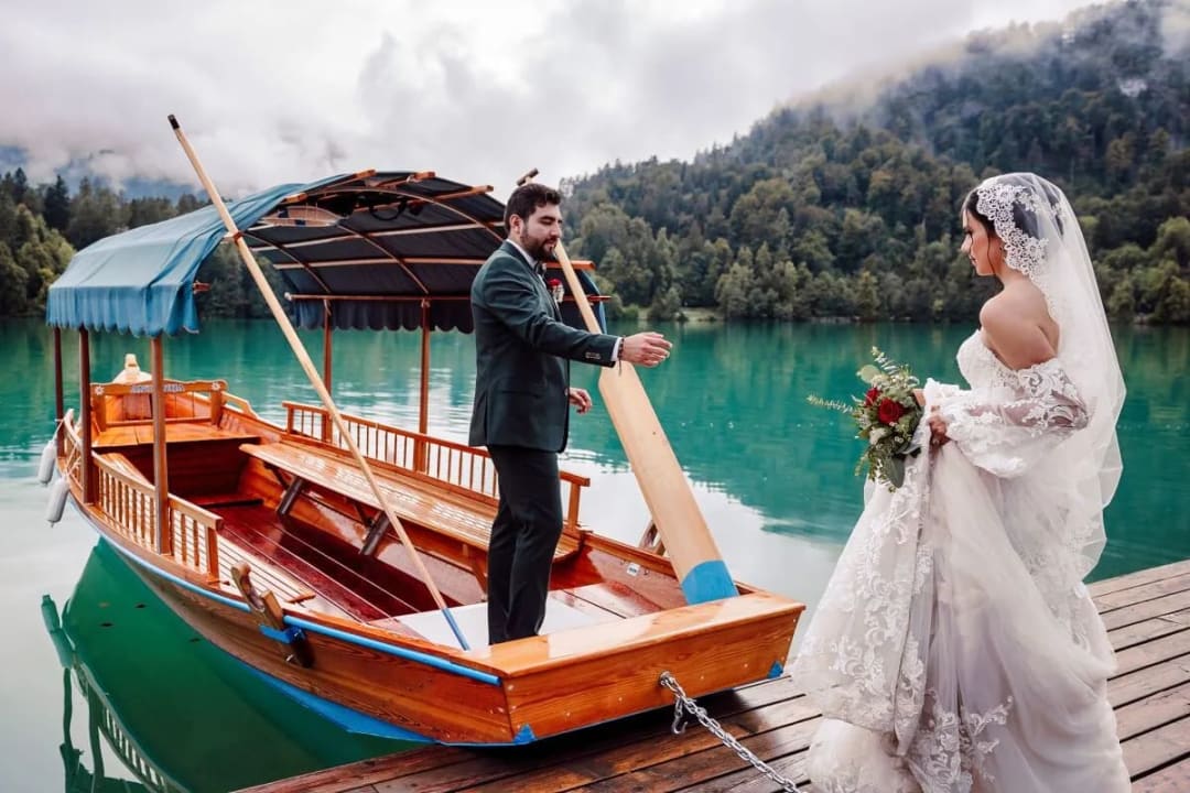 A bride and groom standing by a wooden boat on the calm waters of Lake Bled, surrounded by lush greenery and mountains.