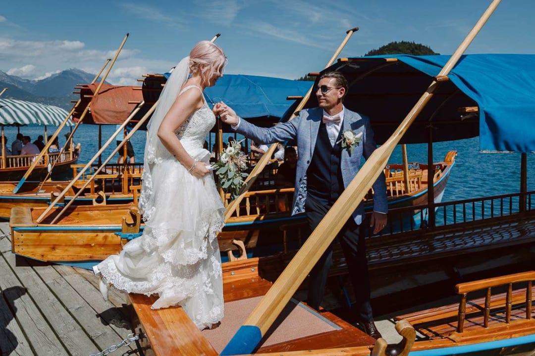 A bride and groom at a boat dock, with the bride stepping onto a boat as the groom offers his hand for support, set against the backdrop of Lake Bled under a clear blue sky.