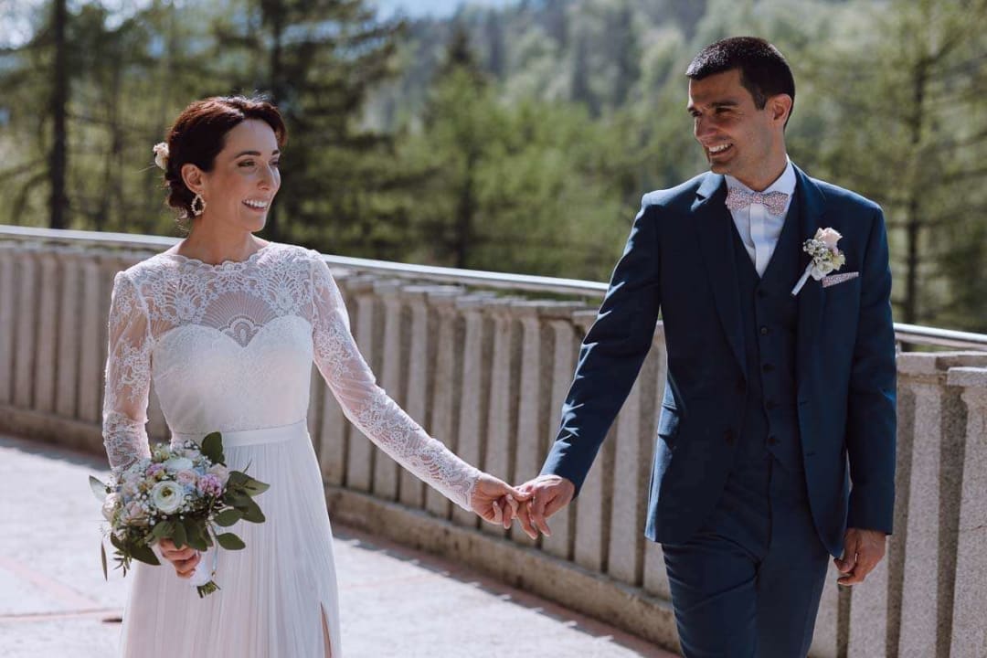 A bride and groom walking hand in hand, smiling at each other in a beautiful outdoor setting.