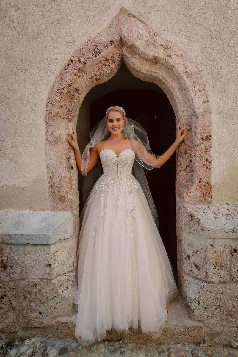A bride in a strapless wedding dress standing in a stone archway, smiling warmly.