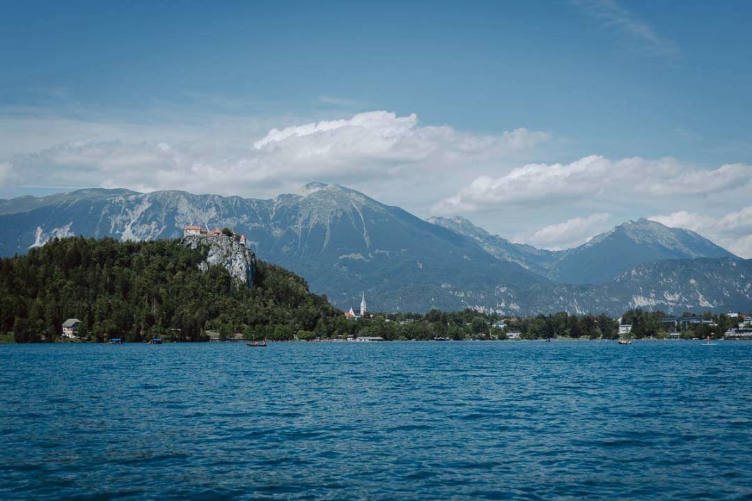 A scenic view of Lake Bled with the castle perched on a hill and mountains in the background under a clear blue sky.