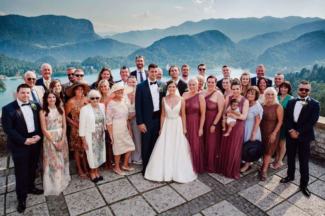A large wedding group photo with the bride and groom surrounded by family and friends, with a scenic view of Lake Bled and the surrounding mountains in the background.