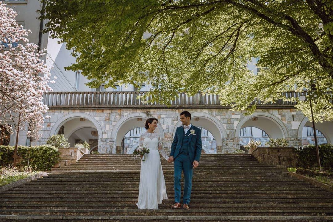 A bride and groom holding hands and smiling at each other while standing on stone steps under leafy trees, with a picturesque building and arches behind them.