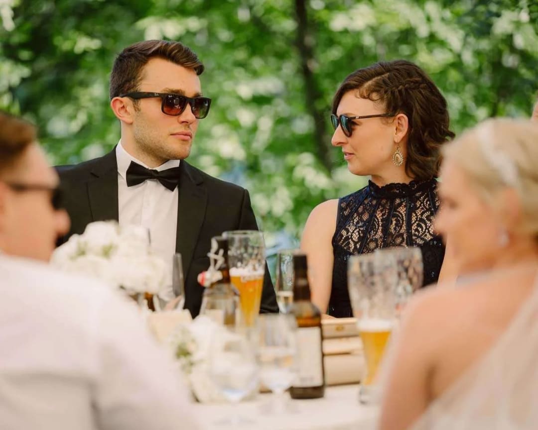 A couple at a wedding reception sitting at a table outdoors, both wearing sunglasses and having a conversation, surrounded by greenery.