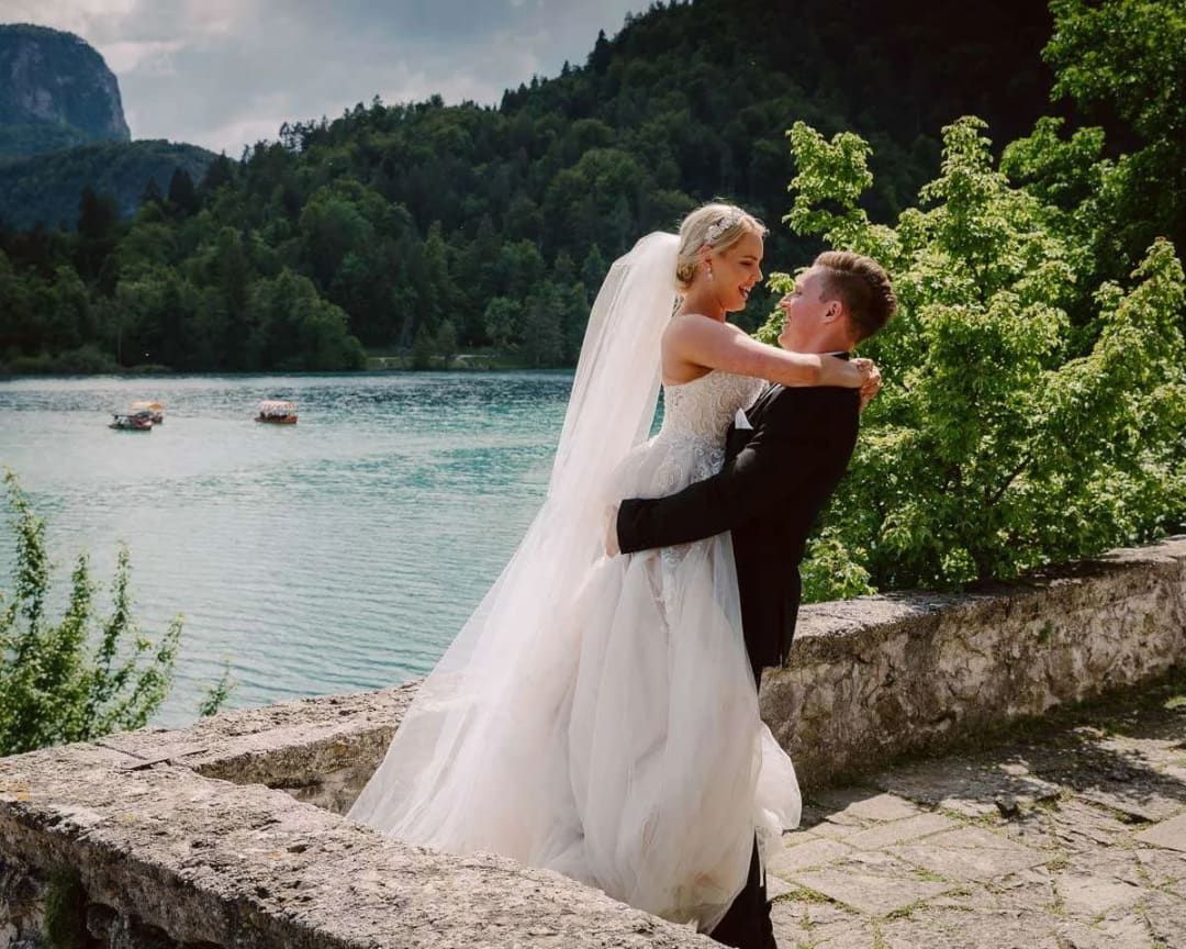 A groom joyfully lifting his bride in his arms with a scenic view of a lake and forested mountains in the background.