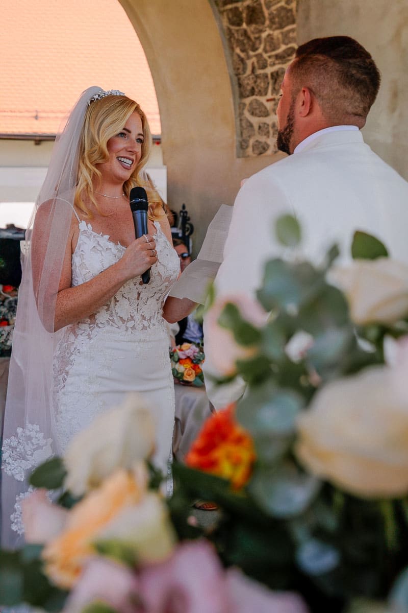 A bride in a lace gown and veil reads vows to the groom during their wedding ceremony, surrounded by colorful flowers.