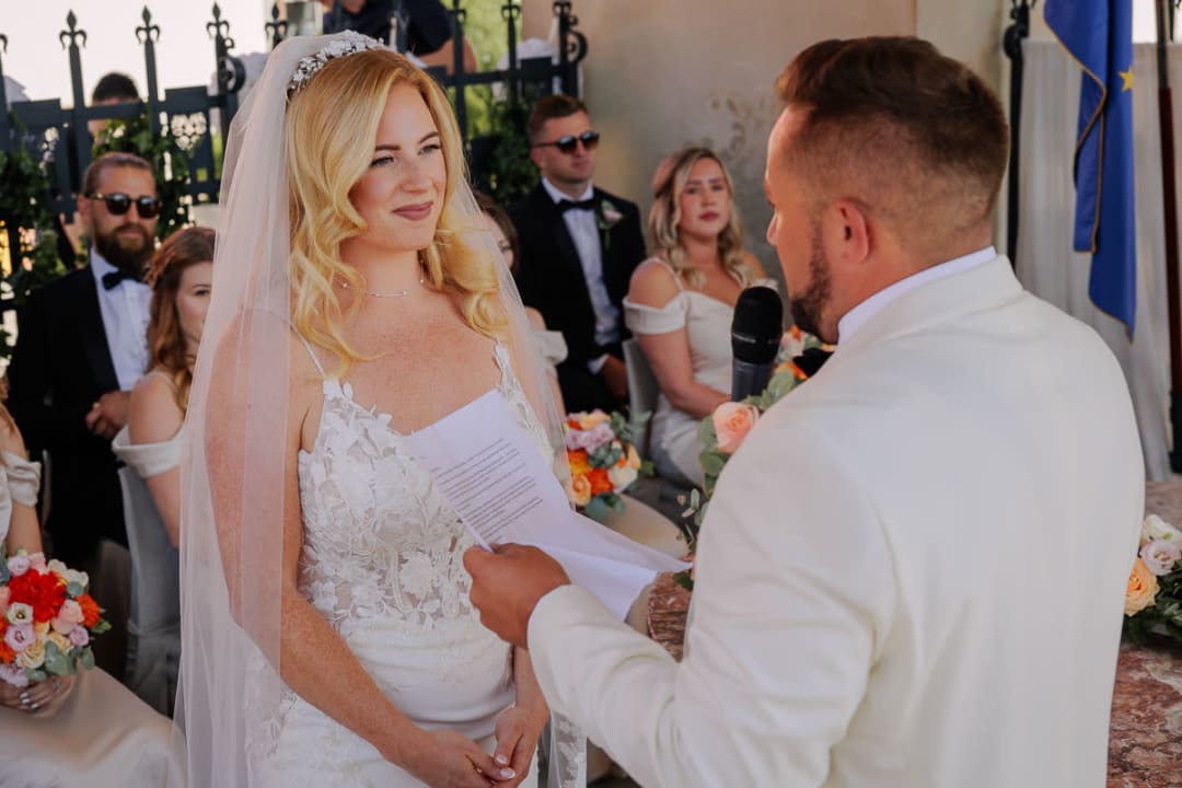 A bride smiles tenderly as the groom reads his vows during their wedding ceremony, with guests seated in the background.