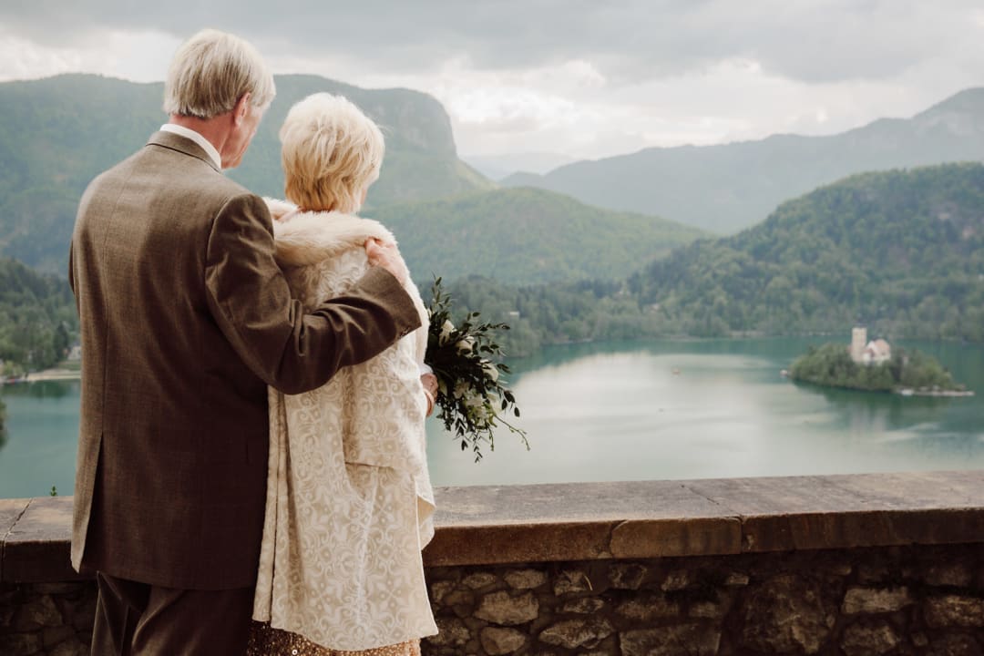 An elderly wedding couple standing together and overlooking Lake Bled from Bled Castle on their wedding day.