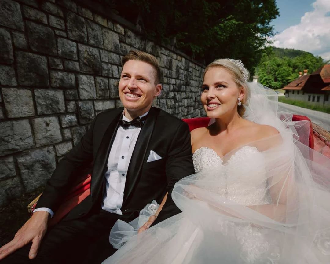 A smiling bride and groom sitting together in a carriage, with a stone wall and lush greenery in the background.