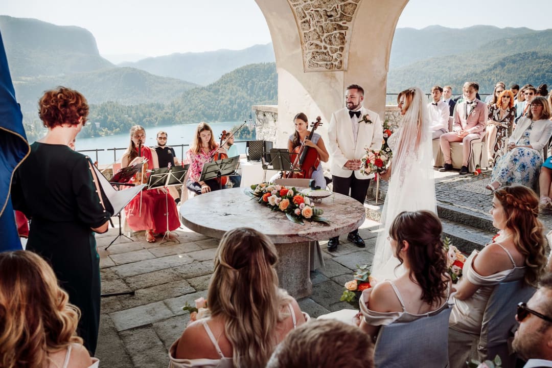 Wide view of a wedding ceremony on the Bled Castle terrace with Lake Bled in the background and live musicians playing.