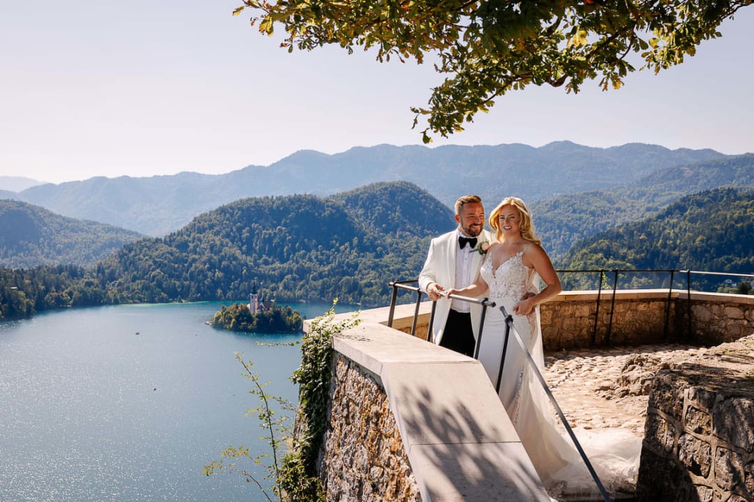 A bride and groom smiling together on a terrace overlooking Lake Bled with its island and surrounding mountains.