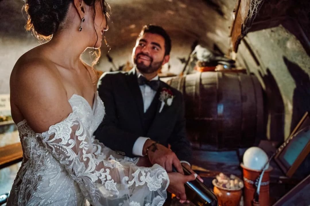A bride and groom smiling at each other in a dimly lit wine cellar, sharing a moment while holding a bottle of wine.