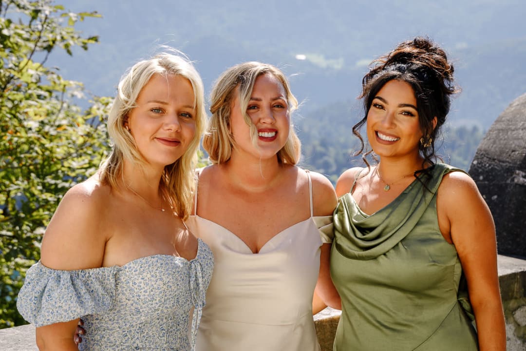 Three women in elegant dresses smiling with a scenic mountain view in the background.