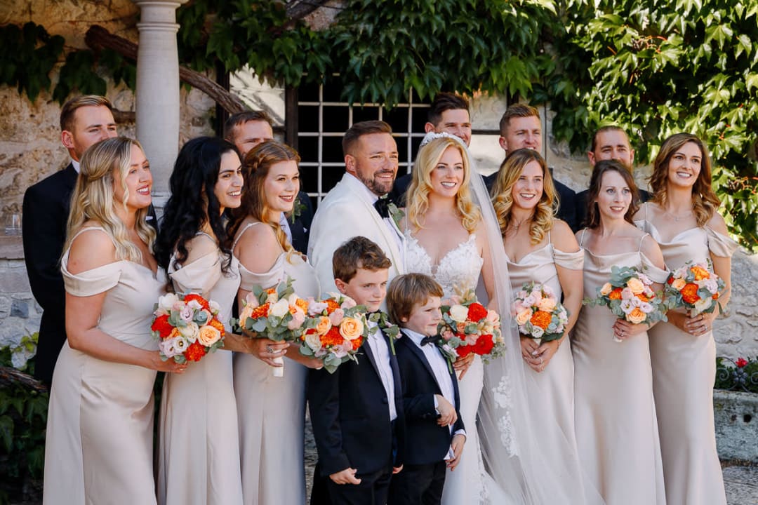 A happy wedding party with the bride and groom standing surrounded by bridesmaids in matching dresses and groomsmen, all smiling and holding vibrant bouquets.