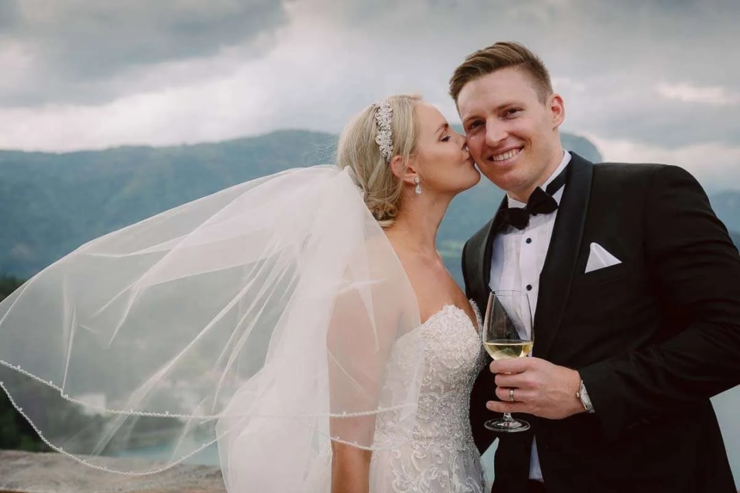 A bride in a white gown kisses the cheek of a smiling groom in a tuxedo, with mountains in the background.