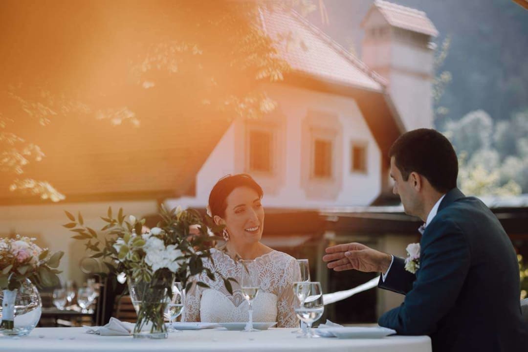A bride and groom sitting at a decorated table, smiling and enjoying their meal outdoors with soft sunlight creating a warm ambiance.