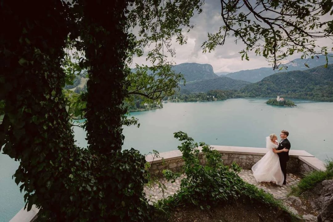 A bride and groom standing together overlooking Lake Bled with a scenic view of the lake and distant mountains.