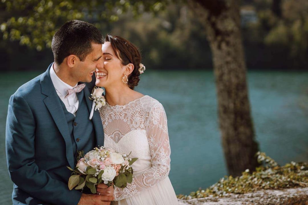A bride and groom smiling warmly at each other while seated by a serene lake.